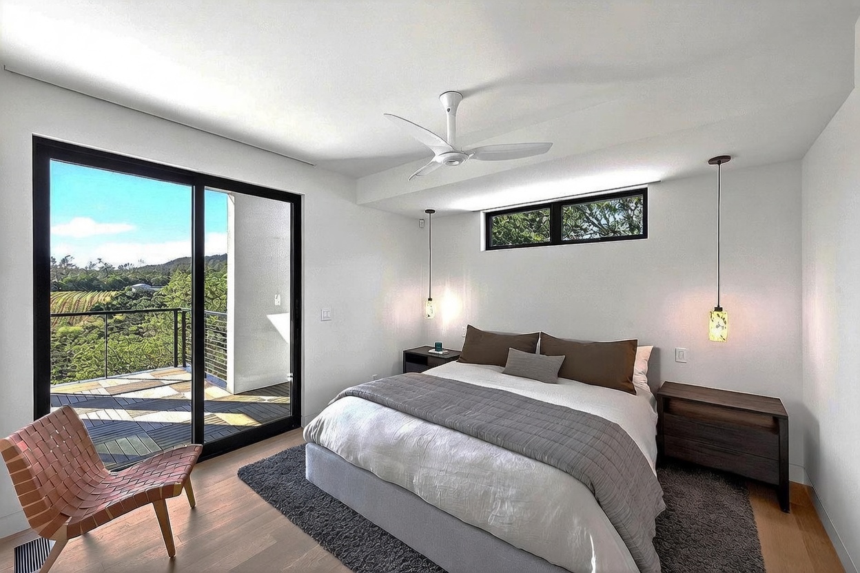 This modern primary bedroom features a minimalist aesthetic with clean white walls, warm wood flooring, and a large sliding glass door that opens onto a private balcony with scenic views. The room is anchored by a plush bed with neutral bedding, flanked by dark wood nightstands and unique pendant lighting, creating a serene and sophisticated atmosphere.