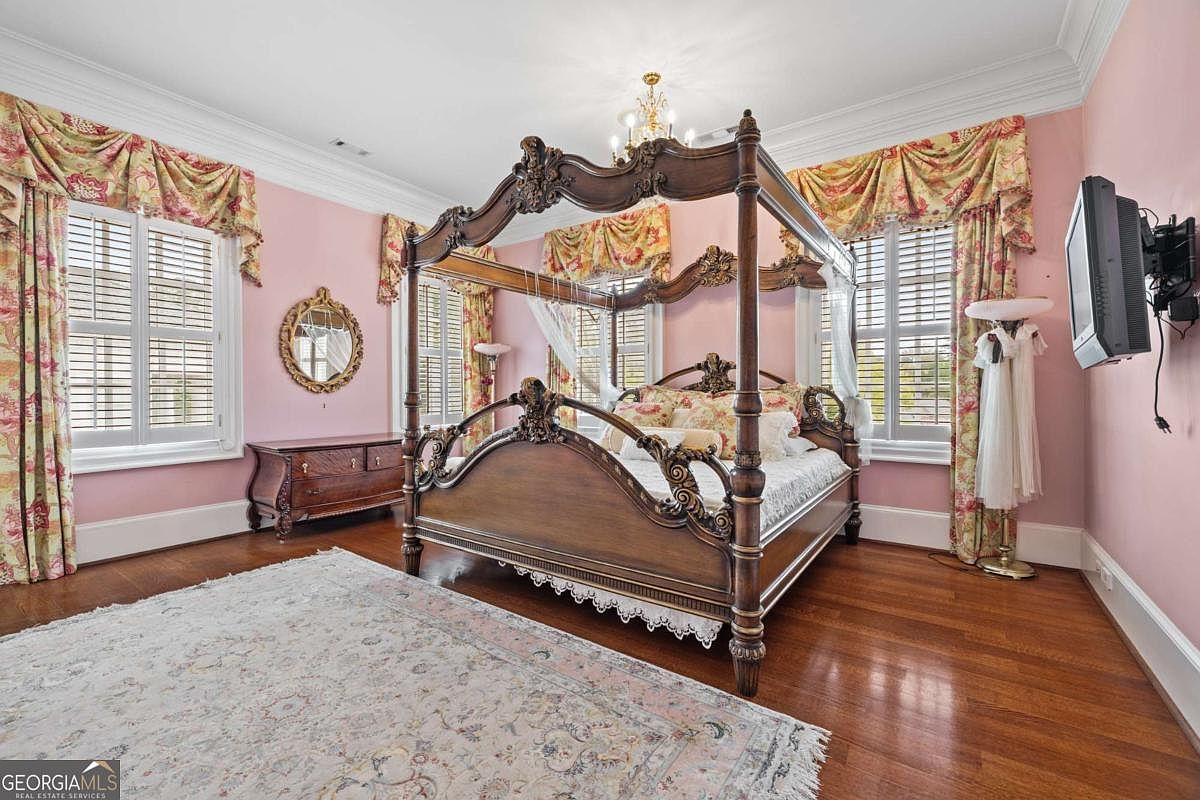 This primary bedroom features a grand, ornate four-poster wooden bed as the central focal point, set against soft pink walls. The room is accented by traditional floral valances over large windows with white shutters, a decorative gold-framed mirror, and a classic wooden dresser. A large, light-toned patterned area rug covers the center of the hardwood floor, contributing to the room's elegant and traditional aesthetic.