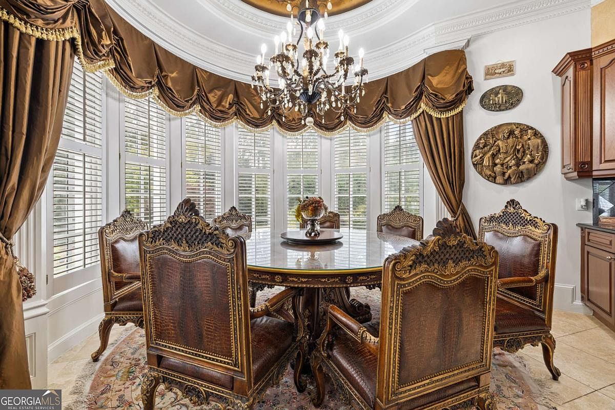 This formal dining room features an ornate, traditional aesthetic centered around a large circular glass-topped table surrounded by high-backed, carved wooden chairs with textured upholstery. A grand crystal chandelier hangs from a recessed ceiling, while a curved wall of tall windows is draped in heavy, draped fabric valances. The room is decorated with wall-mounted sculptural art and sits on a patterned area rug over light-colored tile flooring.