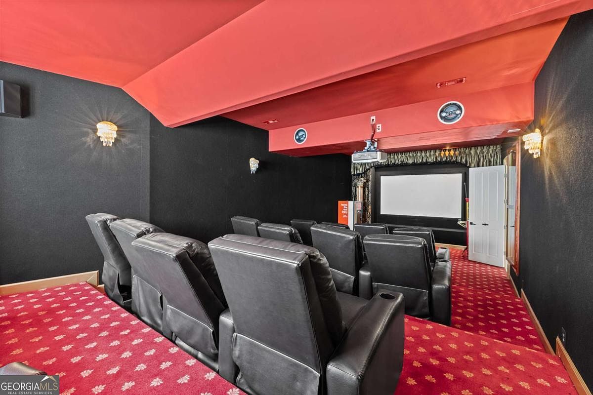 This dedicated home theater room features tiered seating with multiple rows of black leather recliners facing a projector screen. The space is designed with a bold aesthetic, utilizing a vibrant red ceiling and matching red patterned carpeting against dark black walls. A ceiling-mounted projector and wall-mounted sconces provide the lighting, creating an immersive cinematic environment.