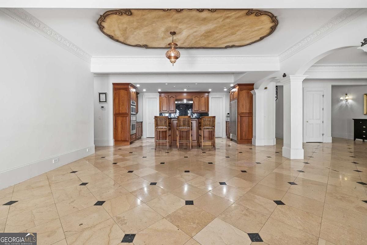 This spacious kitchen features a central island with three wooden bar stools, set against a backdrop of rich-toned cabinetry and dark countertops. The room is anchored by a large, decorative ceiling medallion with a hanging pendant light, and the floor is finished in a light-colored tile with dark accent inlays. The perspective is a wide, eye-level shot looking directly into the kitchen area, highlighting the open-concept layout and the transition into adjacent living spaces.