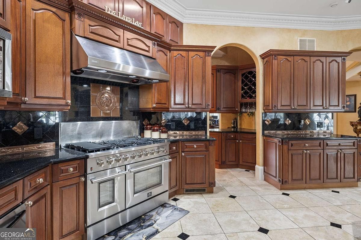 This traditional kitchen features rich, dark-stained wood cabinetry and a professional-grade stainless steel gas range with a matching vent hood. The space is accented by a dark stone backsplash, a decorative medallion inlay, and a built-in wine rack visible through an arched transition. The light-colored tile flooring with dark corner accents provides a classic contrast to the warm wood tones throughout the room.
