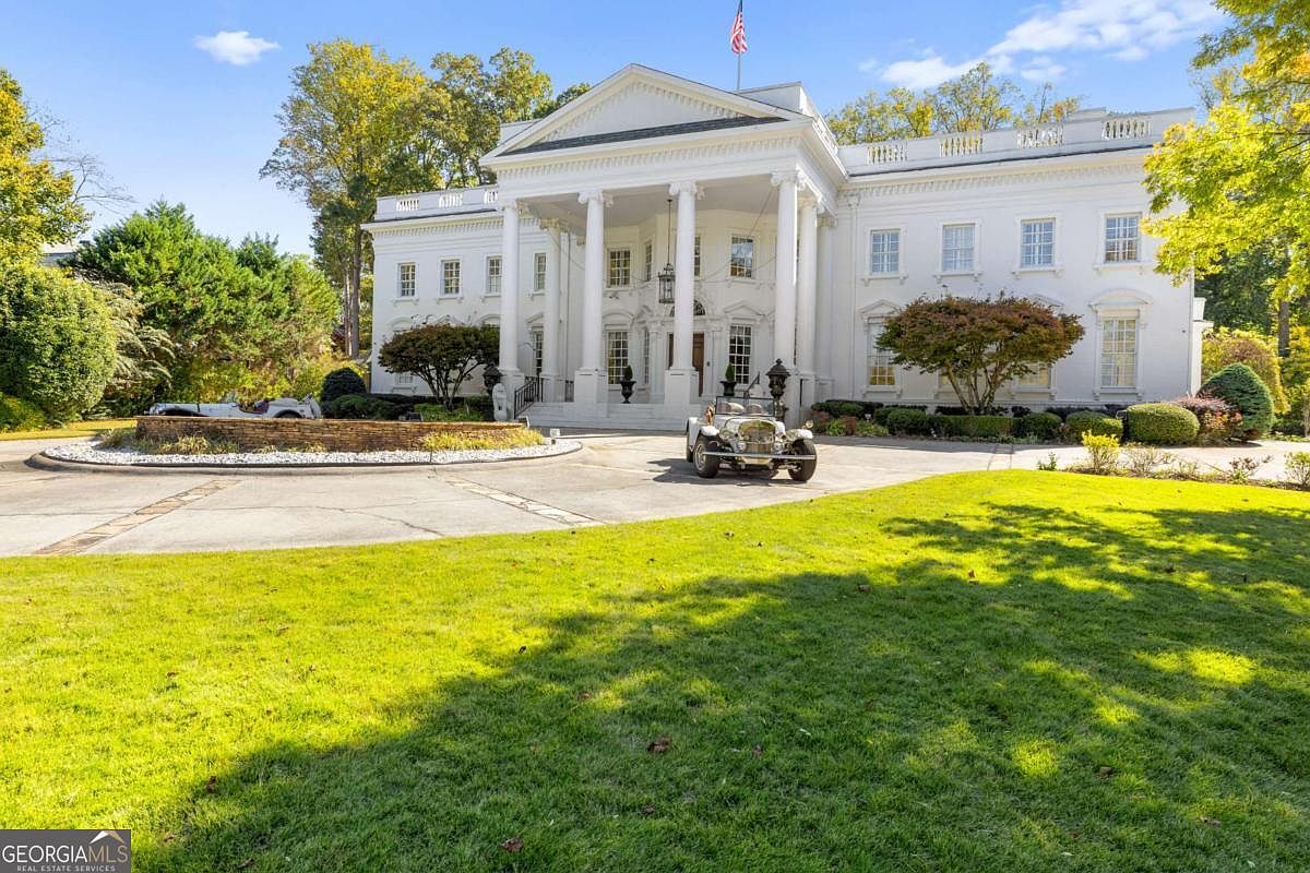 This grand, white neoclassical mansion features a prominent portico with tall columns and a triangular pediment, reminiscent of the White House. A circular driveway leads to the main entrance, where a vintage-style car is parked, and a stone fountain sits in the center of the lawn. The property is surrounded by mature trees and lush green landscaping, creating an imposing and stately curb appeal.