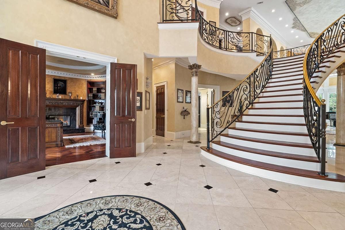 This grand foyer features a sweeping, curved staircase with ornate wrought-iron railings and dark wood treads. The space is finished with light-colored stone flooring with dark accent inlays and warm, textured wall finishes. To the left, double doors open into a study with a fireplace and built-in shelving, creating a sense of luxury and architectural depth.