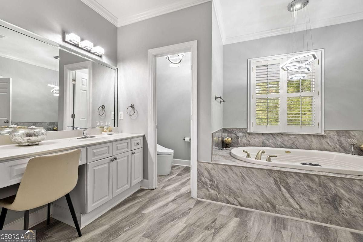 This primary bathroom features a neutral color palette with light gray walls and wood-look flooring. The space includes a long vanity with a makeup station, a separate water closet, and a large soaking tub encased in stone-patterned tile. A modern light fixture hangs above the tub, and the room is brightened by natural light coming through shuttered windows.