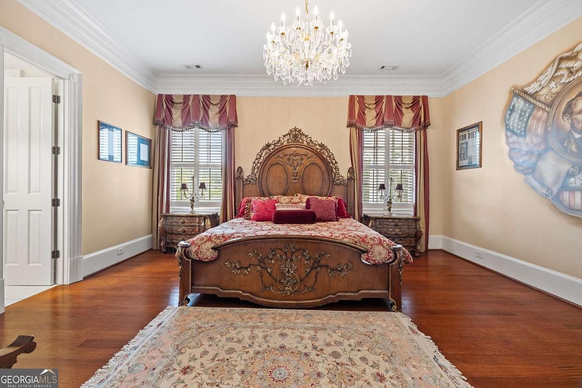 This primary bedroom features a grand, ornate wooden bed frame as the central focal point, set against a soft-toned wall. The room is illuminated by a large, multi-tiered crystal chandelier and includes two windows dressed with elegant valances and shutters. A patterned area rug rests on the polished wood flooring, while a large decorative wall plaque adds a unique, historical touch to the space.