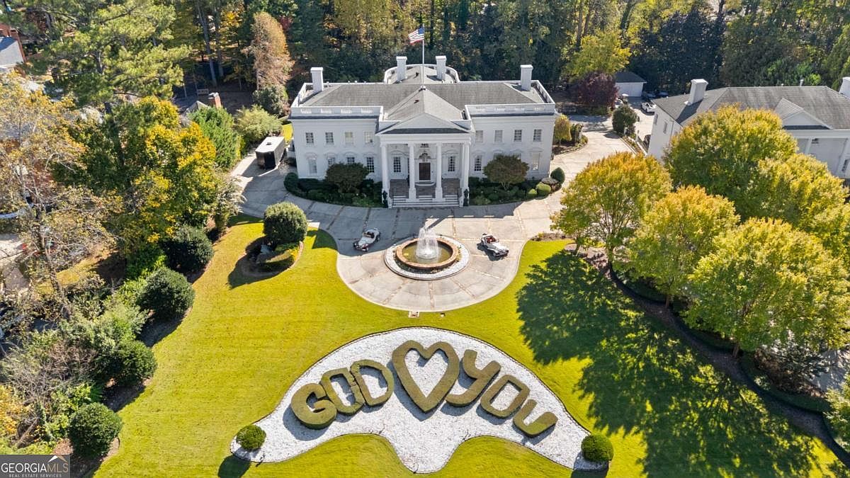 This high-angle aerial view captures a grand, white mansion designed with a striking resemblance to the White House, complete with a central portico and a prominent American flag. The expansive front lawn features a circular driveway with a central fountain and a large, decorative landscaping installation that spells out 'GOD ❤️ YOU'. The property is surrounded by mature trees and lush greenery, creating a stately and private estate atmosphere.