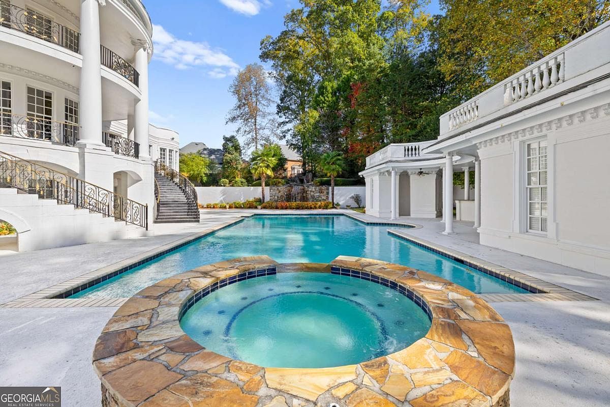 This expansive outdoor living area features a large, rectangular swimming pool paired with a circular stone-clad spa in the foreground. The property showcases a grand, multi-story white mansion with curved architectural details, elegant wrought-iron railings, and a stone staircase leading to the pool deck. A covered poolside pavilion with columns and a balustrade roofline adds a classic, luxurious touch to the serene backyard setting.