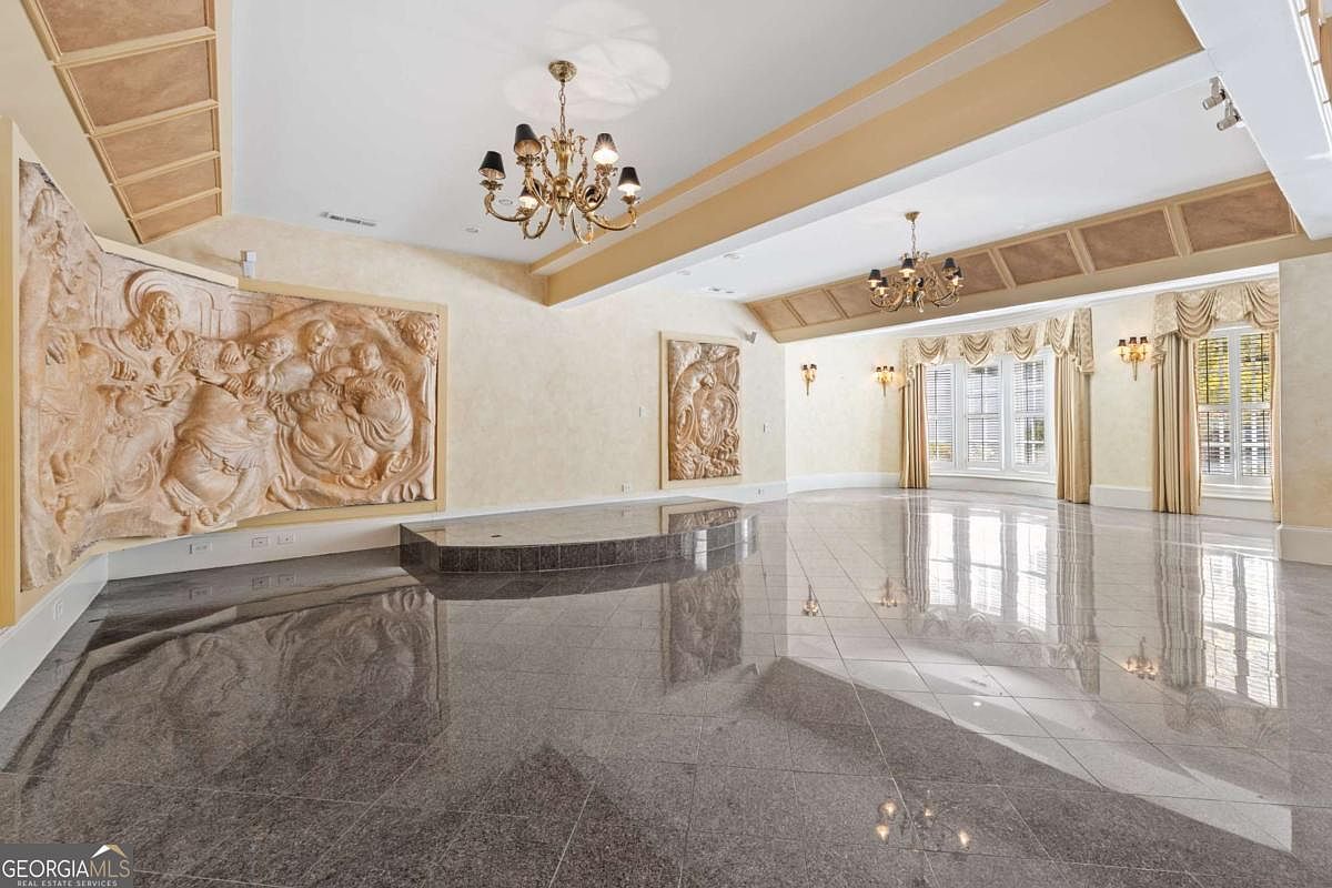 This expansive, open-concept living space features highly polished, reflective stone flooring and a raised platform area. The room is distinguished by large, intricate relief sculptures mounted on the walls and is illuminated by two ornate, multi-tiered chandeliers. Large windows with elegant drapery allow natural light to fill the room, creating a grand and formal atmosphere.