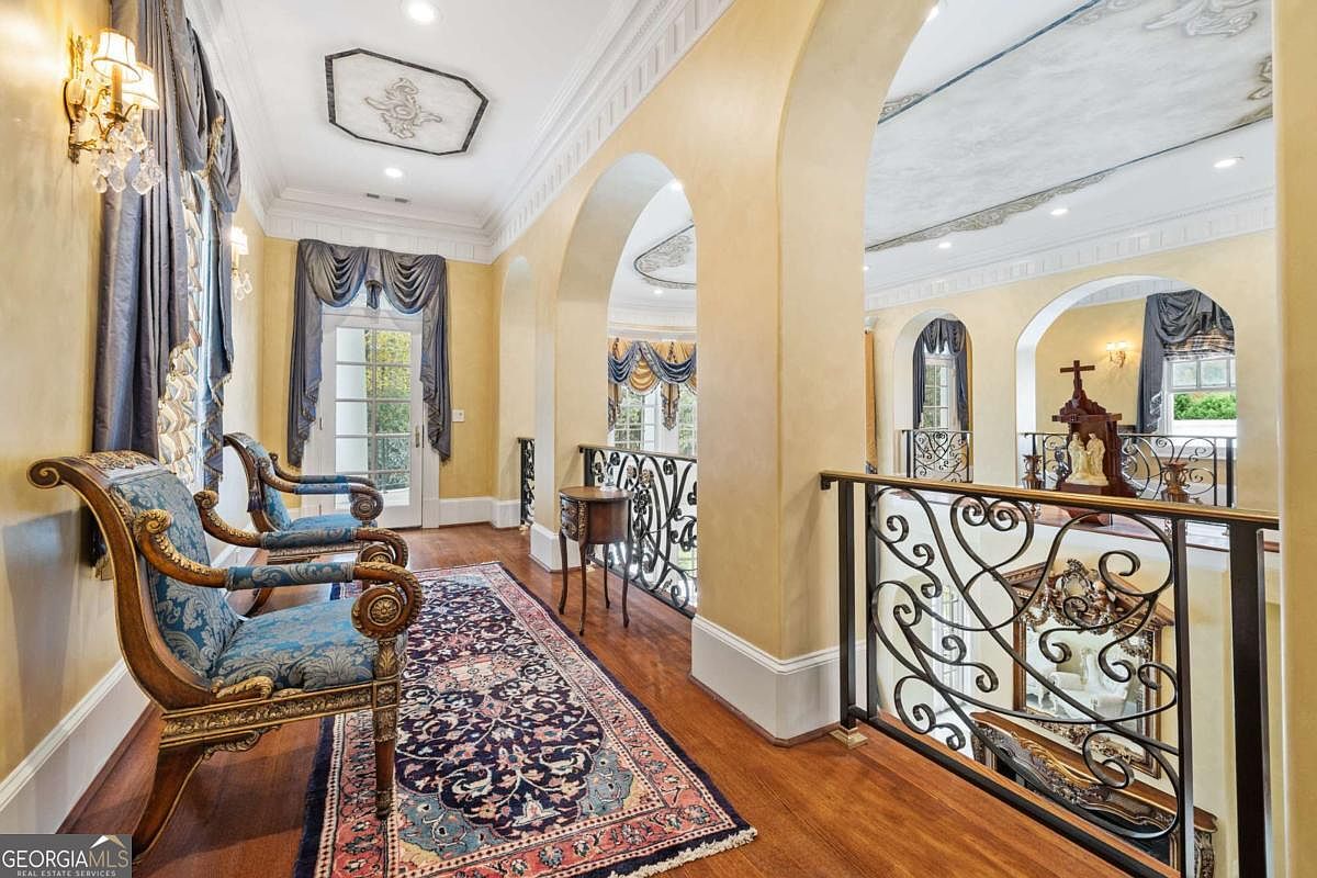 This elegant upper-level hallway features a classic, ornate design with arched openings and a decorative wrought-iron railing overlooking the floor below. The space is furnished with two traditional blue upholstered chairs and a small wooden side table, all set upon a detailed patterned rug over hardwood floors. The warm yellow walls, intricate crown molding, and soft lighting create a sophisticated and formal atmosphere.