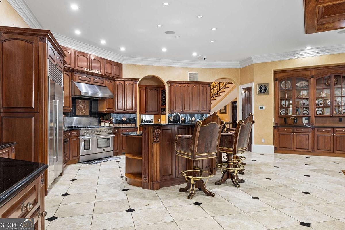 This spacious, traditional-style kitchen features rich, dark wood cabinetry and a central island with bar seating. The room is anchored by a large stainless steel range and a built-in refrigerator, complemented by light-colored tile flooring with dark decorative accents. A large, glass-fronted display cabinet adds a formal touch, while the open layout provides a clear view toward a staircase in the background.