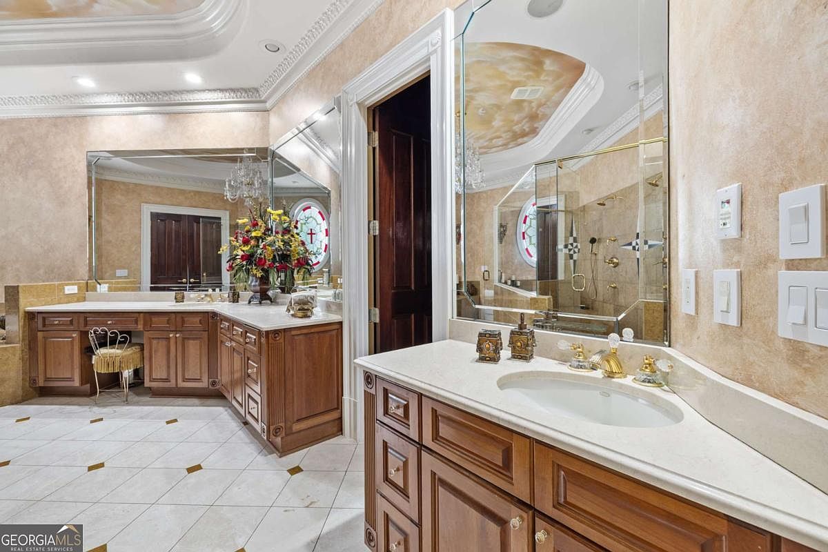This spacious primary bathroom features dual vanities with wood cabinetry and light-toned stone countertops, complemented by large wall-to-wall mirrors. The room is accented with decorative faux-finish walls, a tiled floor with inset diamond patterns, and a glass-enclosed shower area. A vanity stool and a floral arrangement add a touch of elegance to this traditional-style bathroom.