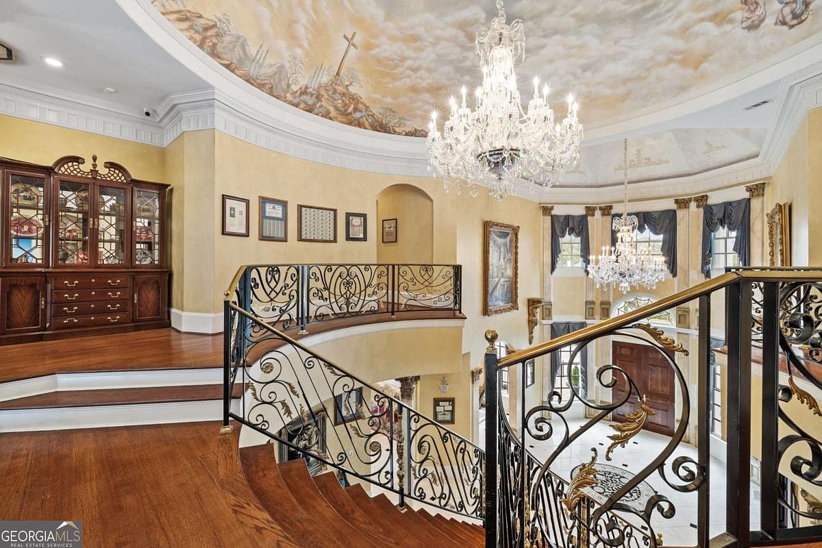 This grand foyer features a sweeping staircase with intricate wrought-iron railings that lead to an upper landing. The ceiling is adorned with a detailed mural of clouds and a cross, illuminated by a large, ornate crystal chandelier. A dark wood hutch sits on the upper level, while the space is finished with warm-toned walls and polished wood flooring, creating an opulent and formal atmosphere.