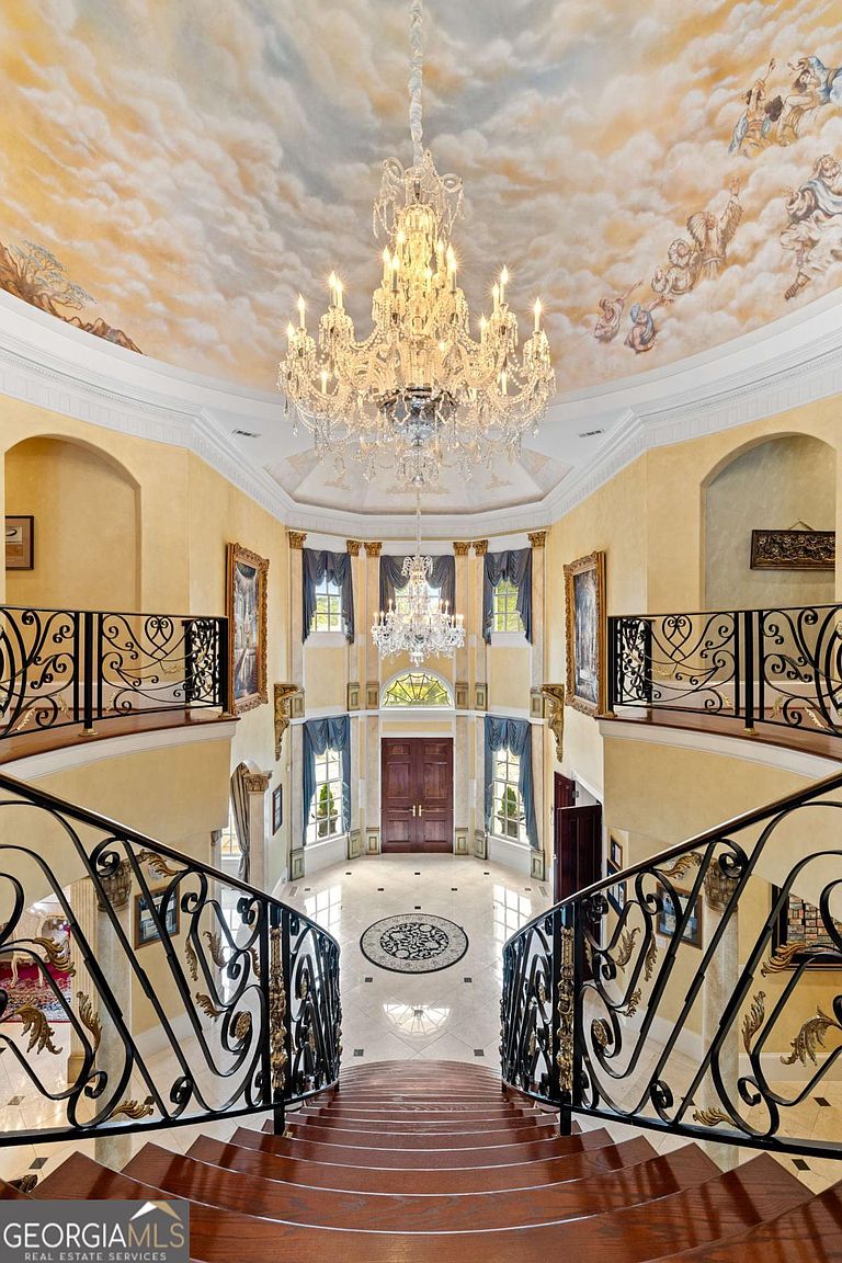 This grand foyer features a sweeping staircase with ornate wrought-iron railings that lead down to a polished stone floor with a central medallion. Above, a massive crystal chandelier hangs from a domed ceiling adorned with a detailed mural of clouds and figures. The space is framed by tall windows with blue drapery and symmetrical balconies, creating an opulent and formal atmosphere.