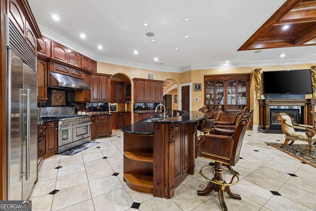 This expansive, traditional-style kitchen features rich wood cabinetry, a large central island with a dark stone countertop, and a professional-grade stainless steel range. The space flows seamlessly into an adjacent living area, which includes a fireplace with a decorative mantel and a wall-mounted television. The room is finished with light-colored tile flooring with dark accent inlays, creating a sophisticated and cohesive open-concept layout.