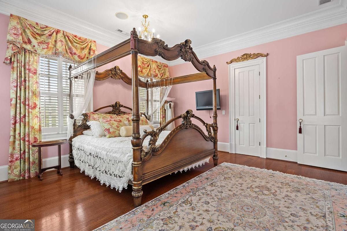 This primary bedroom features a grand, ornate four-poster wooden bed as the central focal point, set against soft pink walls. The room is accented by traditional floral valances over shuttered windows and a large, intricate area rug covering the polished wood floors. A television is mounted on the wall, and two white doors provide access to other parts of the home, creating a classic and elegant atmosphere.