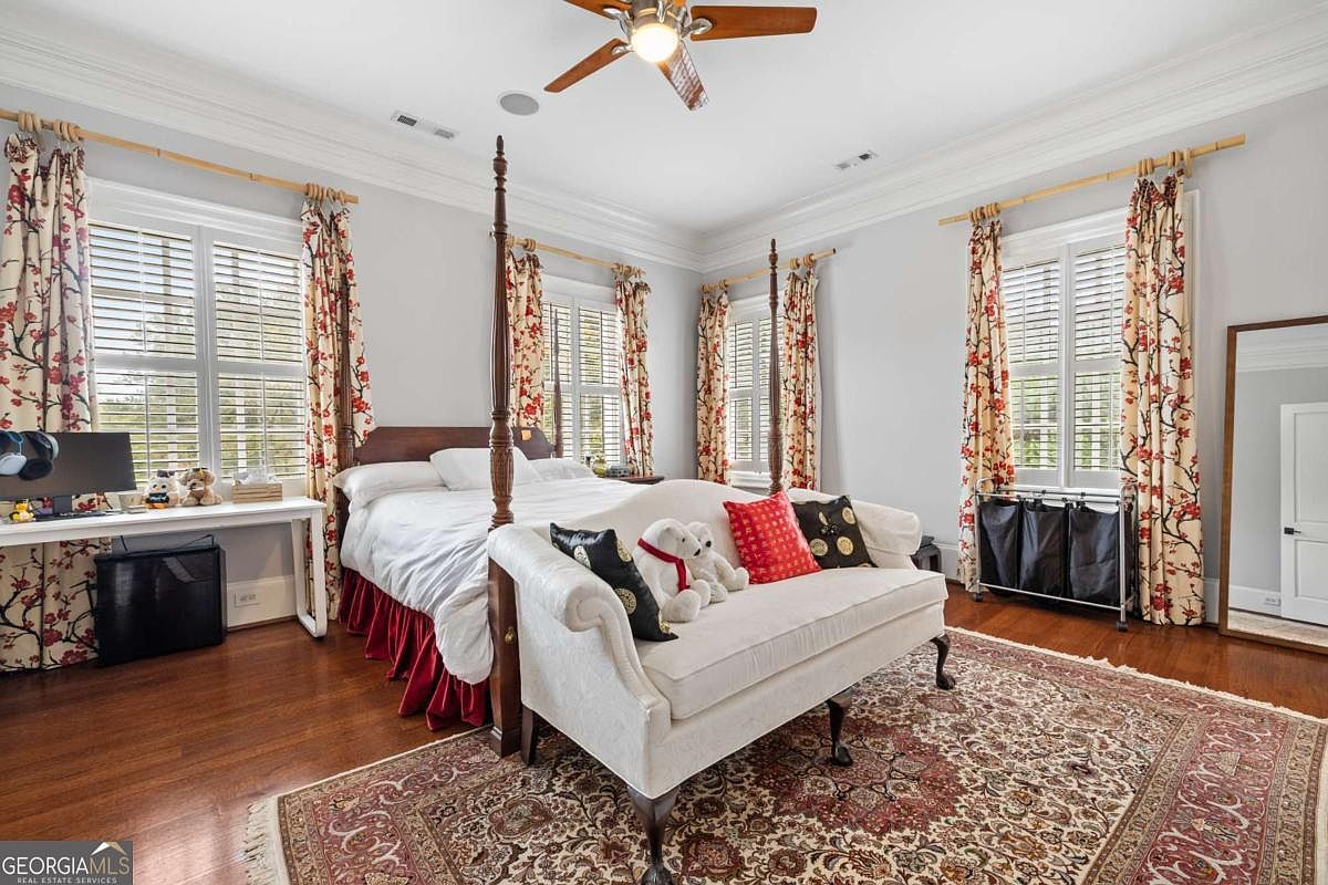 This spacious primary bedroom features hardwood flooring, a four-poster bed, and a cream-colored upholstered bench at the foot of the bed. The room is accented with floral curtains on the windows, a patterned area rug, and a ceiling fan with integrated lighting. A desk area is positioned to the left, and a large floor mirror stands against the wall on the right, creating a bright and traditional living space.
