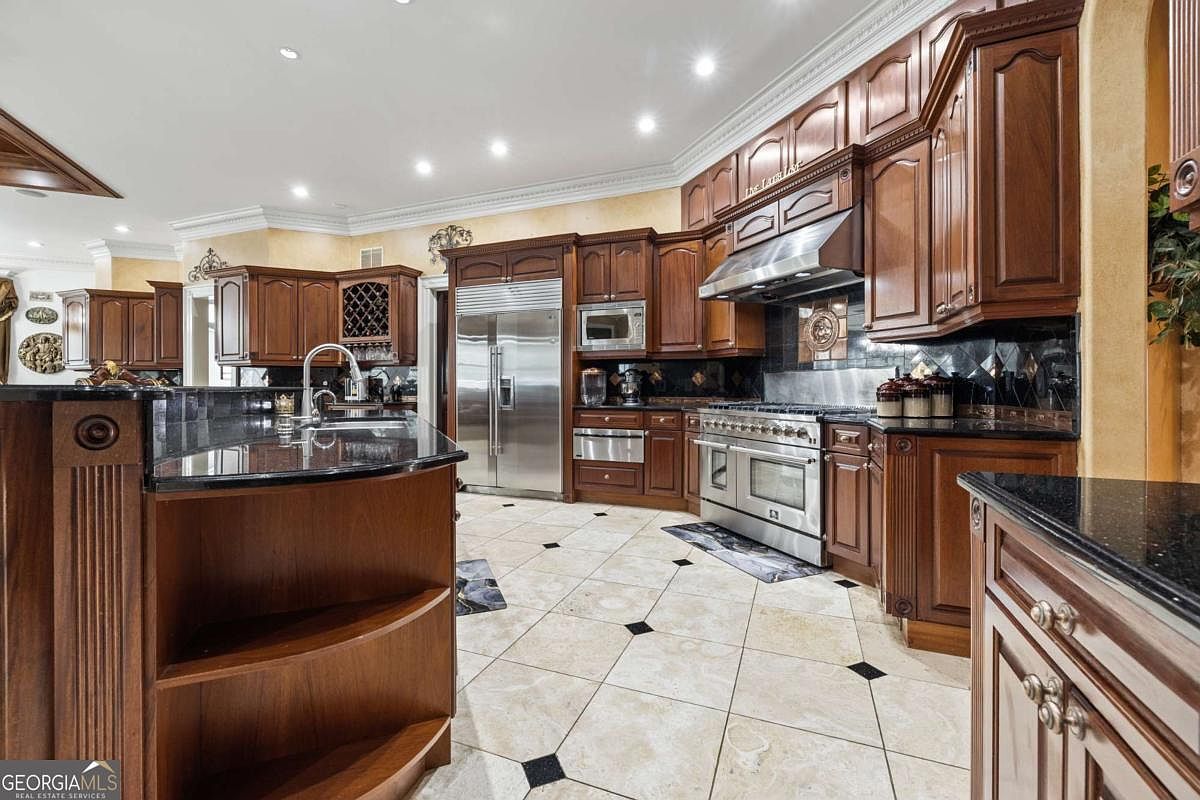 This spacious kitchen features rich, dark-toned cabinetry, a large central island with a curved end, and polished dark stone countertops. The room is equipped with professional-grade stainless steel appliances, including a large range and a built-in refrigerator, set against a backdrop of patterned tile flooring with dark accent inlays. The perspective captures the expansive layout, highlighting the ample storage and the functional, high-end design of the culinary space.