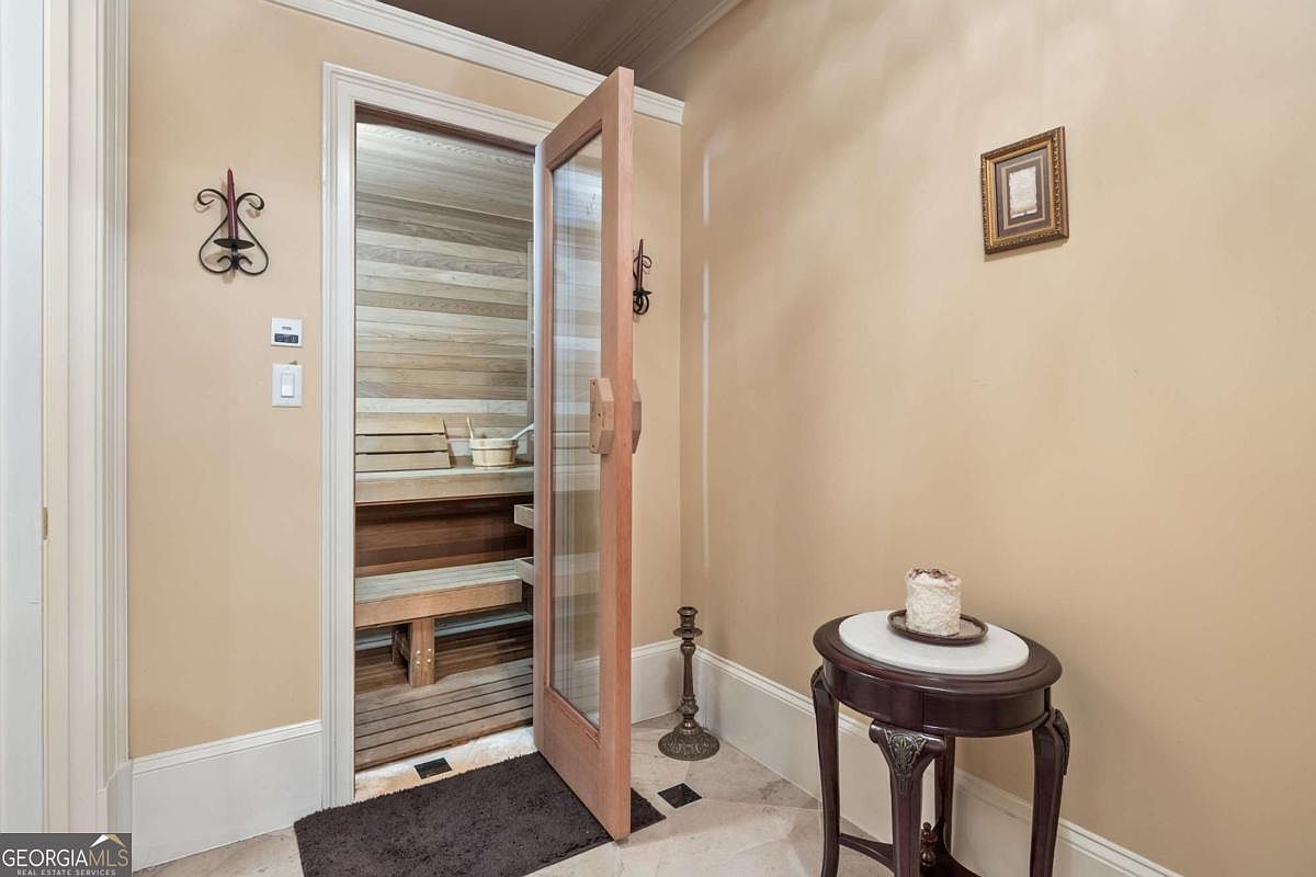 This interior space features a dedicated sauna room accessed through a glass-paneled wooden door. The sauna interior is lined with light-toned wood slats and includes built-in benches and a wooden bucket. Outside the sauna, a small round side table with a decorative object sits on a light-colored tile floor, complemented by a metal candle holder and a wall-mounted decorative iron piece.
