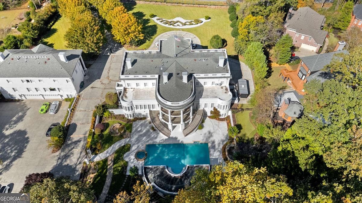 This high-angle aerial view captures a grand, white mansion designed with a striking resemblance to the White House, complete with a central curved portico and symmetrical wings. The property features an expansive paved driveway, a large swimming pool with an integrated spa, and meticulously landscaped grounds surrounded by mature trees. The perspective highlights the impressive scale of the estate, its formal architectural layout, and the private, wooded setting.