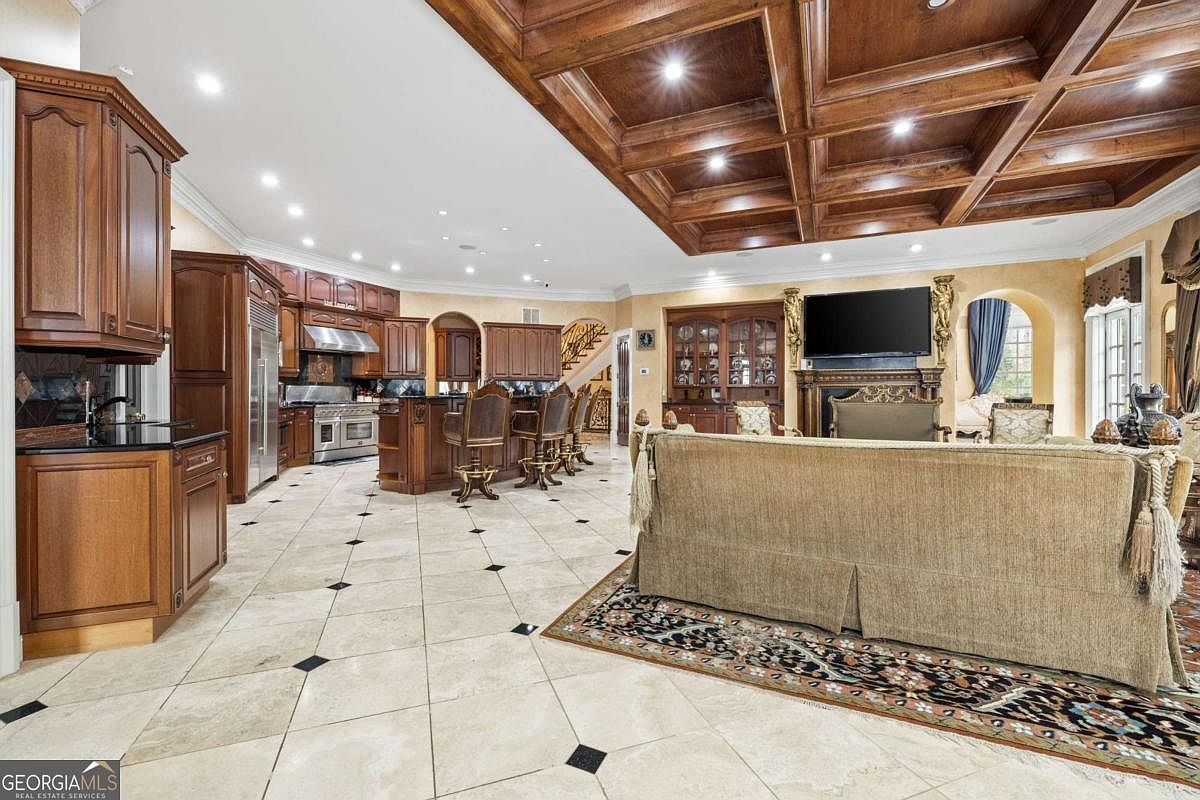 This expansive open-concept space features a large kitchen with dark wood cabinetry, a central island with bar seating, and light-colored tile flooring with dark accent inlays. The living area is anchored by a large sofa facing a television mounted above a traditional fireplace, all set beneath a dramatic coffered wood ceiling. The perspective captures the seamless flow between the culinary workspace and the comfortable lounge, highlighting the home's grand scale and traditional design aesthetic.