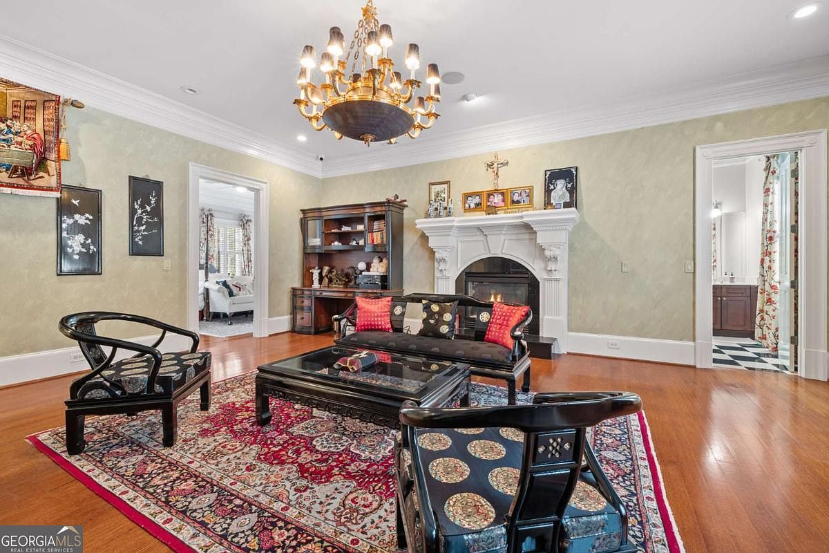 This elegant living room features a formal seating arrangement centered around a fireplace with a decorative white mantel. The space is adorned with a large, ornate chandelier, a patterned area rug, and dark wood furniture with Asian-inspired design elements. The walls are finished in a soft, textured paint, and the room opens into adjacent spaces, creating a sophisticated and traditional atmosphere.