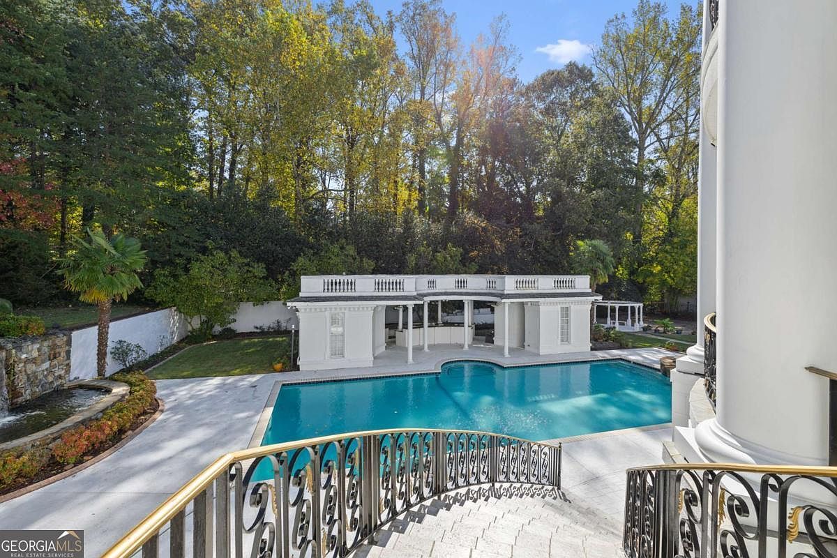 This elevated perspective captures a luxurious backyard featuring a large, curved swimming pool set against a white, classical-style pool house with columns and a balustrade. A stone-clad water feature sits to the left, while a decorative metal railing in the foreground frames the view of the manicured lawn and dense, mature trees in the background. The scene conveys an elegant, resort-like atmosphere perfect for outdoor entertaining.