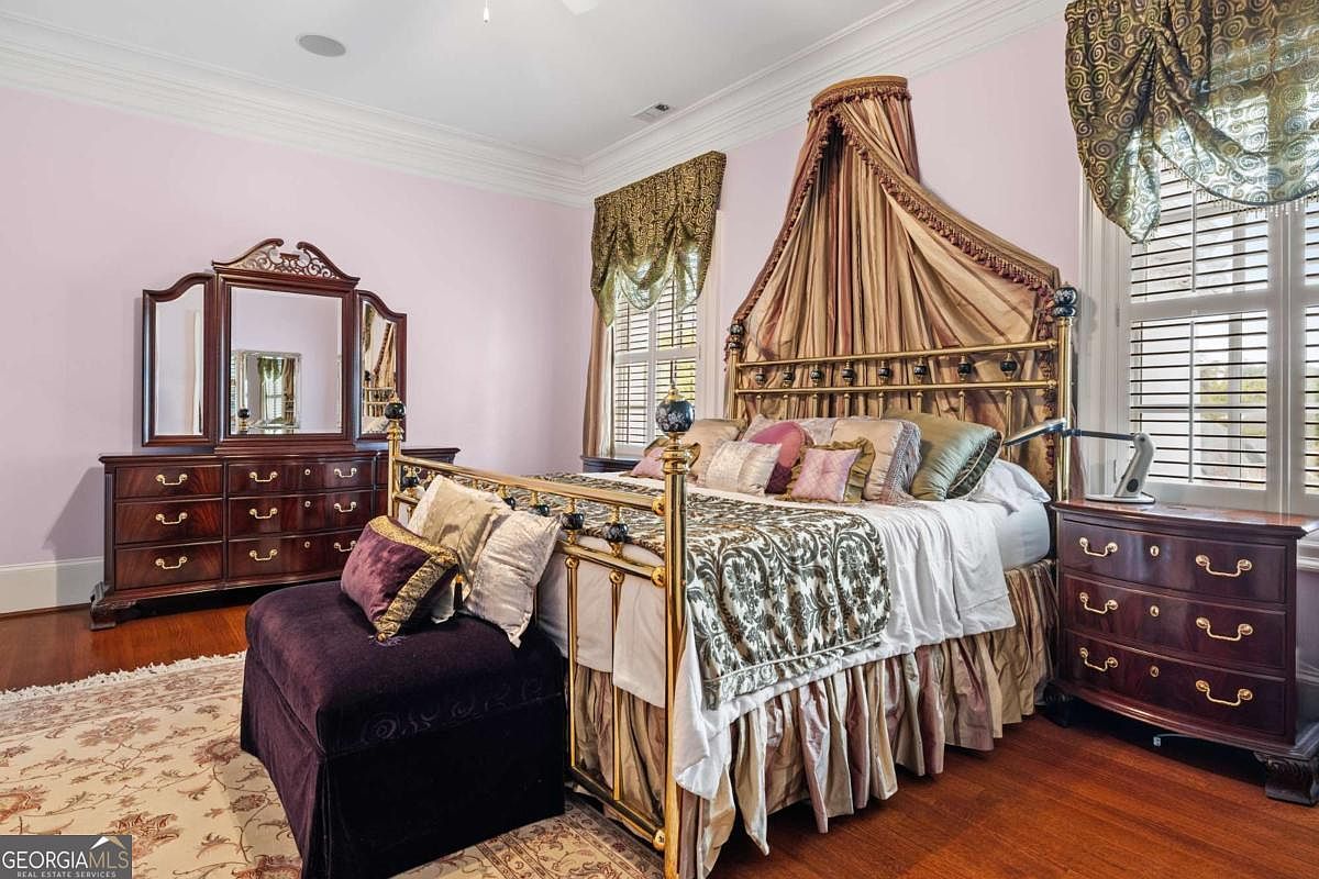 This primary bedroom features a grand, ornate metal bed frame with a draped canopy and a matching dark wood dresser with a tri-fold mirror. The room is decorated with soft pink walls, patterned window treatments, and a plush purple bench at the foot of the bed. The space is anchored by hardwood flooring and a large area rug, creating a traditional and opulent atmosphere.