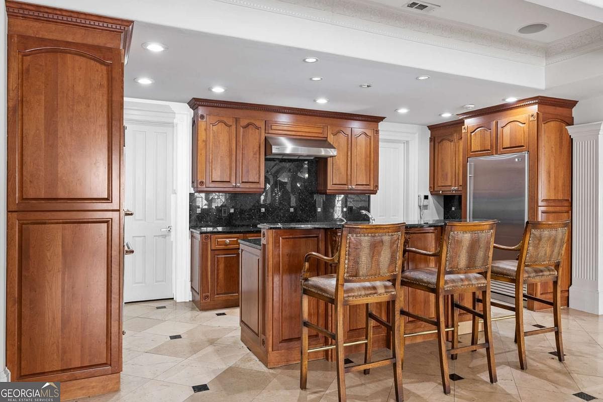 This kitchen features warm-toned wood cabinetry, dark stone countertops, and a central island with three bar stools. The space is illuminated by recessed lighting and includes a stainless steel refrigerator and a built-in range hood. The flooring consists of light-colored tiles with dark accent inlays, creating a traditional and cohesive aesthetic.