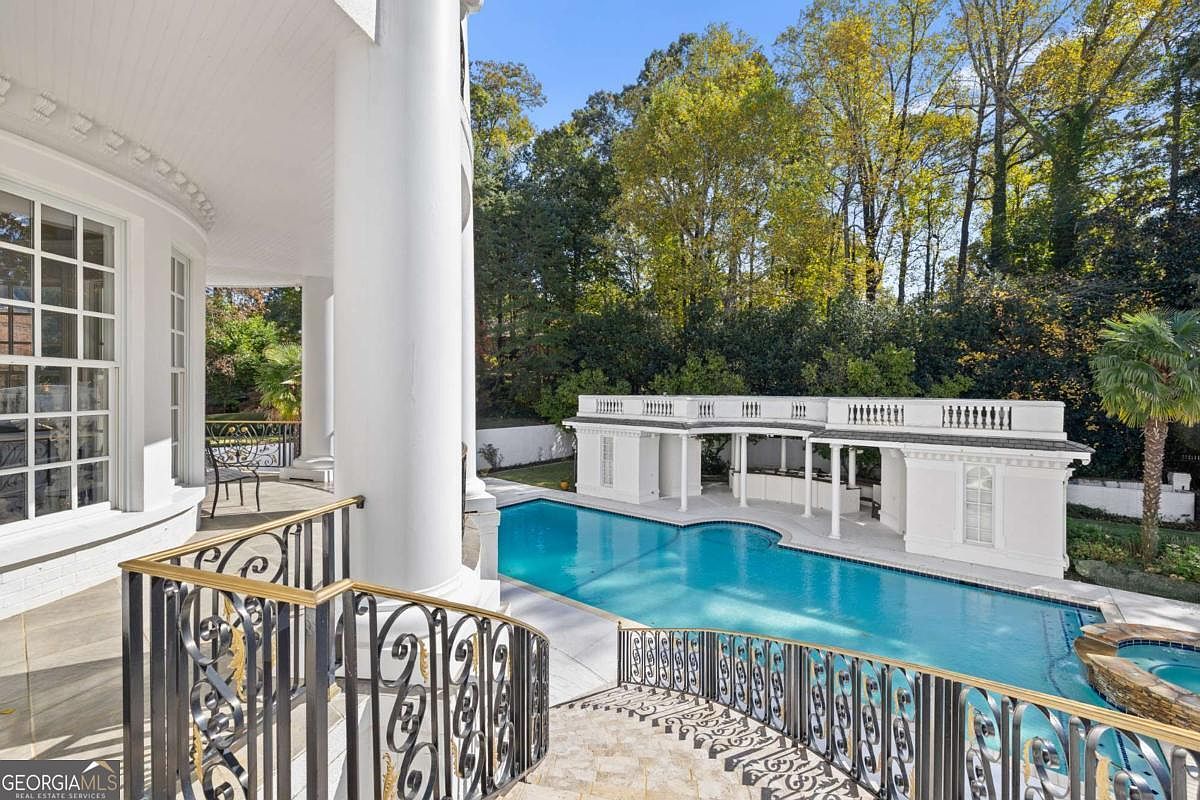 This elevated perspective captures a luxurious outdoor living space featuring a curved swimming pool and an adjacent spa. A grand, white-columned terrace with ornate black metal railings overlooks the pool area, which is complemented by a classic pool house with a balustrade roofline. The scene is framed by lush, mature trees, creating a private and elegant backyard retreat.