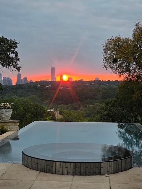 This image showcases a luxurious infinity pool with an integrated spa, offering a stunning view of the city skyline at sunset. The pool's design seamlessly blends with the natural landscape, creating a serene and upscale ambiance. The foreground features the pool's edge and spa, while the background captures the vibrant colors of the setting sun over the cityscape.