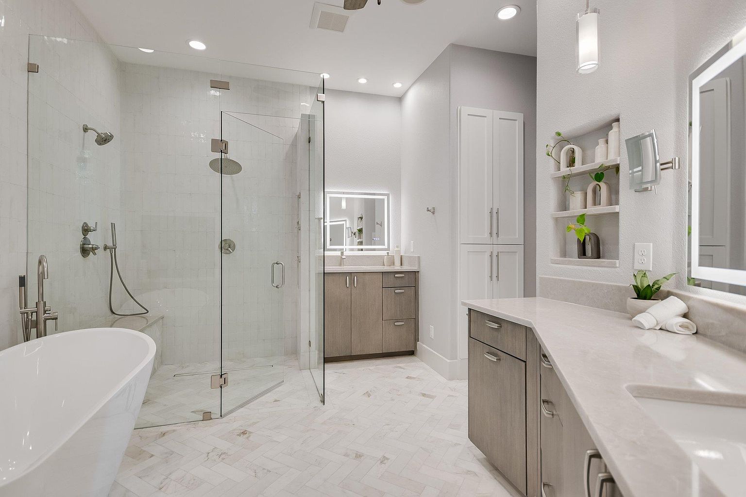 This is a luxurious primary bathroom featuring a freestanding bathtub next to a glass-enclosed shower with a built-in bench. The double vanity has light countertops and modern cabinetry, complemented by a large, illuminated mirror. The flooring is a stylish herringbone pattern, and the overall design is clean, bright, and contemporary.