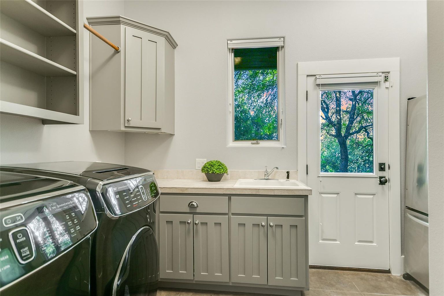 This is a well-organized laundry room featuring a front-loading washer and dryer set, gray cabinetry with a countertop, and a sink. A window provides natural light, and a door leads to the outside. The room appears clean and functional, ideal for a modern home.