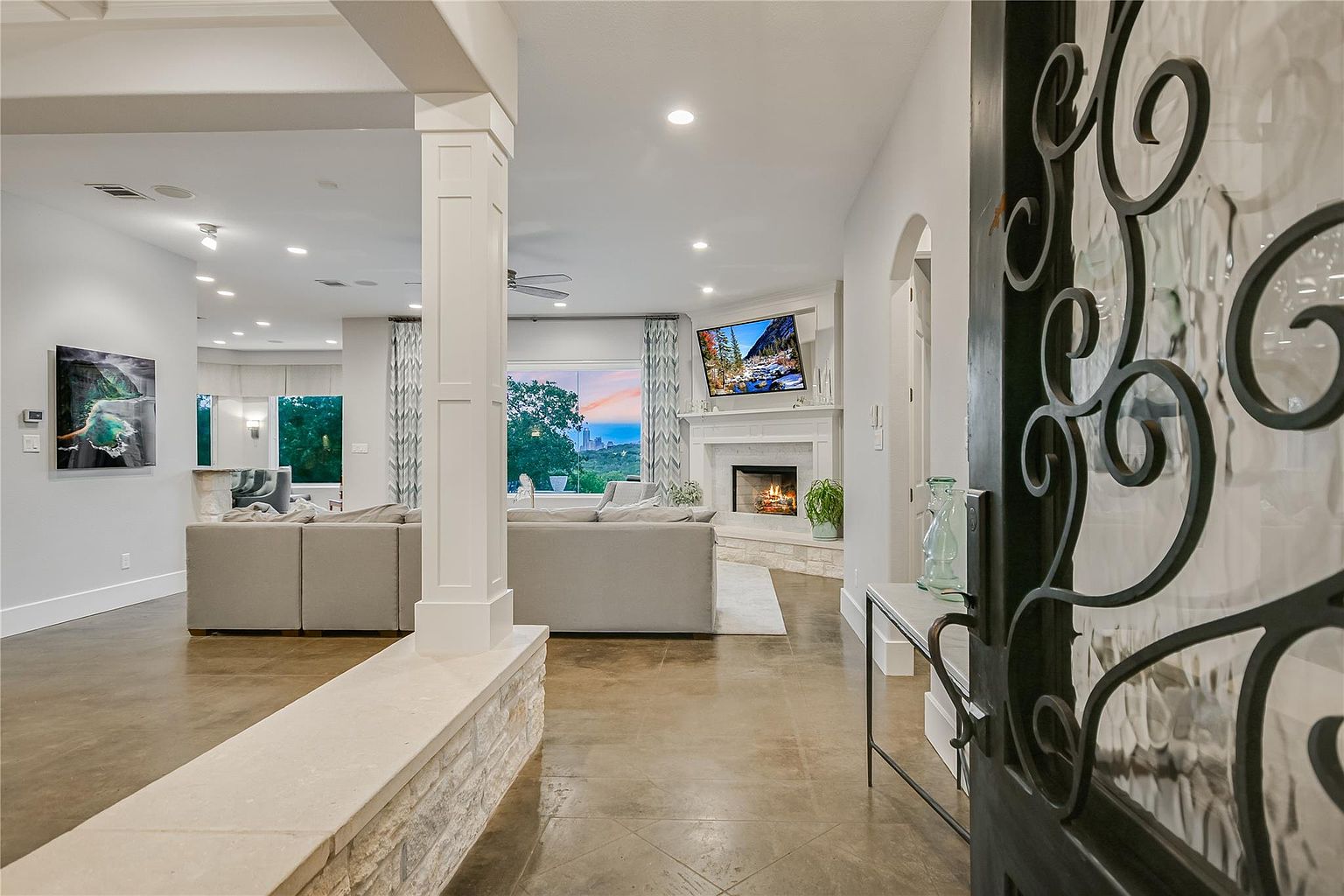 This inviting living room features an open floor plan with a neutral color palette, creating a bright and airy atmosphere. A stone accent wall and decorative columns add architectural interest, while a fireplace with a mounted television serves as a focal point. The room is furnished with comfortable, modern sofas, offering a welcoming space for relaxation and entertainment.