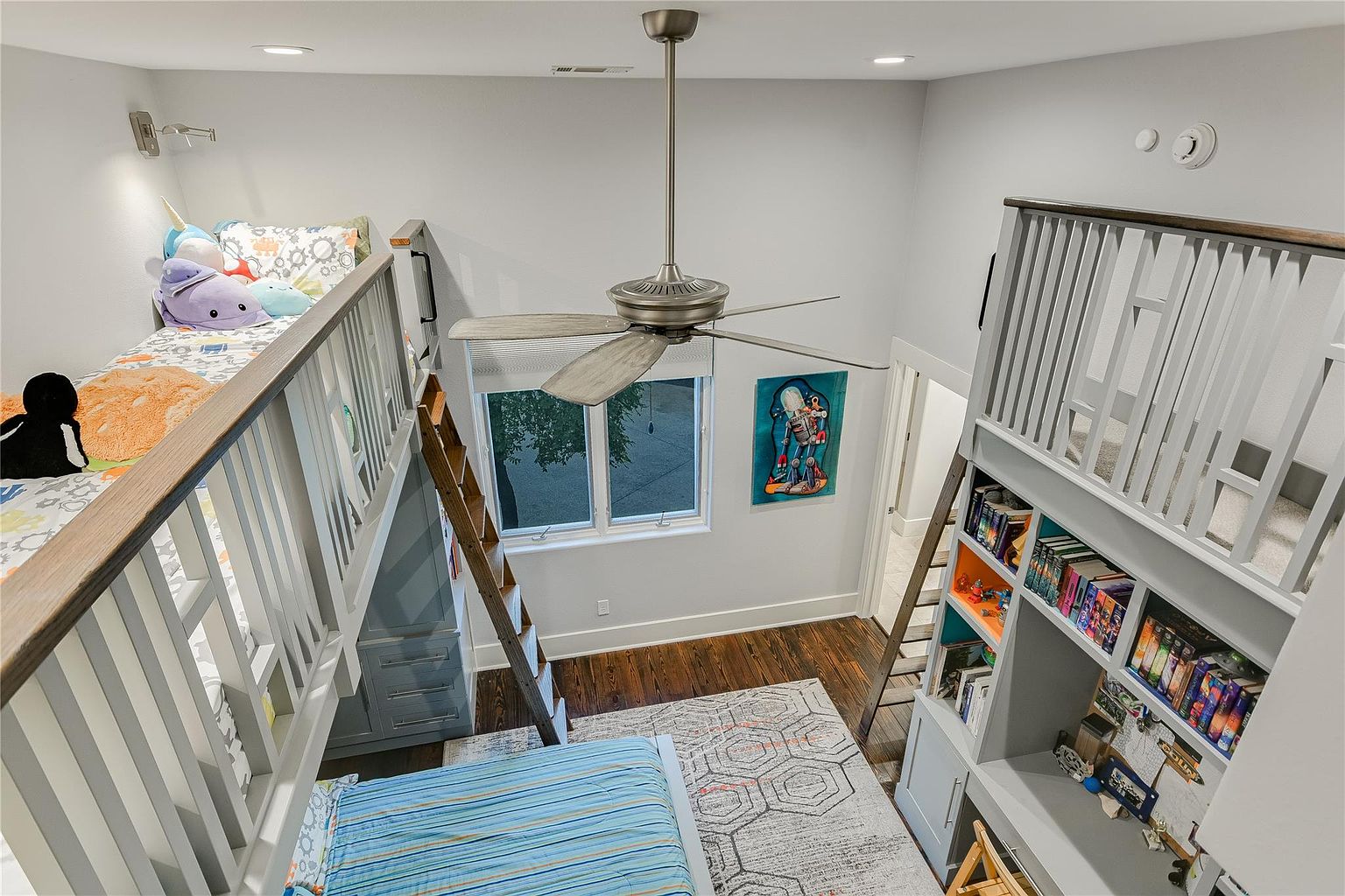 This is a unique bedroom featuring custom-built bunk beds with integrated storage and a study area. The room has a playful and functional design, with a ceiling fan, a window providing natural light, and a cozy rug on the hardwood floor. The overall impression is a well-organized and inviting space for children or guests.