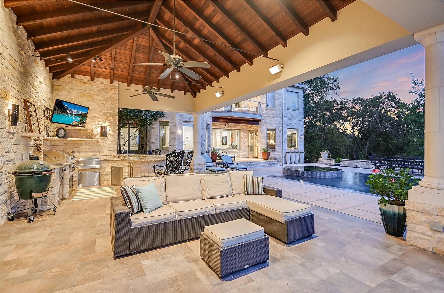This is a luxurious outdoor patio area featuring a comfortable wicker sectional sofa with plush cushions and an ottoman. The space includes a built-in outdoor kitchen with a grill, a television, and ceiling fans for comfort. The patio overlooks a pool and spa area, with lush landscaping and a beautiful home exterior in the background, creating an inviting and upscale outdoor living space.