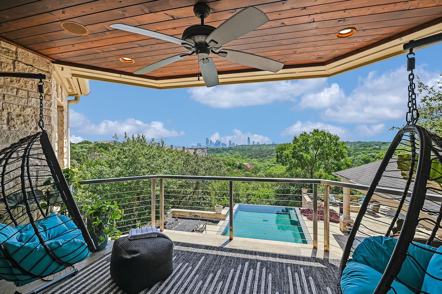 This image showcases a luxurious outdoor balcony or deck area, featuring comfortable hanging chairs with blue cushions and a stylish striped rug. A modern railing provides safety and an unobstructed view of the surrounding lush greenery and a swimming pool below. In the distance, a city skyline adds a touch of urban sophistication, creating a serene and upscale outdoor living space.