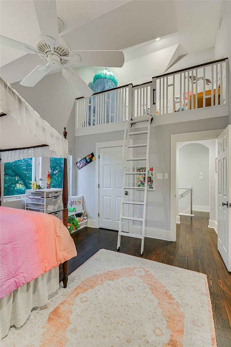 This bedroom features a unique loft area accessible by a white ladder, adding a playful element to the space. The room includes a four-poster bed with a pink and orange ombre comforter, a ceiling fan, and dark hardwood floors complemented by a light-colored rug with a peach-colored pattern. Natural light streams in through the windows, creating a bright and inviting atmosphere.