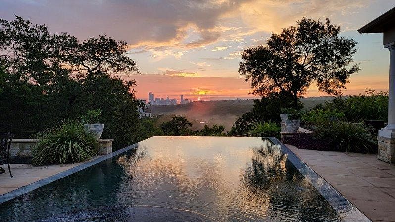 This image showcases a luxurious infinity pool overlooking a stunning cityscape at sunset. The pool's dark water reflects the vibrant sky, creating a serene and elegant atmosphere. Lush greenery surrounds the pool area, enhancing the sense of privacy and tranquility, making it an ideal outdoor space for relaxation and entertainment.