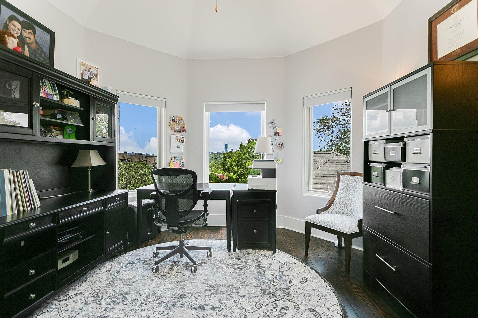 This is an interior shot of a well-organized home office. The room features dark wood furniture, including a large desk, a hutch, and a filing cabinet, complemented by a patterned round rug. Natural light floods the space through multiple windows, offering a view of the outdoors, while a comfortable office chair and an accent chair provide seating.
