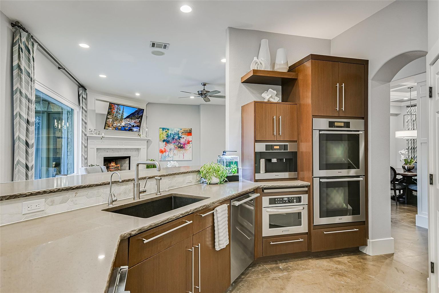 This is a well-lit kitchen featuring wood cabinets, stainless steel appliances, and a dark sink. The countertops are light-colored and the backsplash is a decorative tile. The kitchen opens to a living area with a fireplace and a television, creating an open-concept living space.