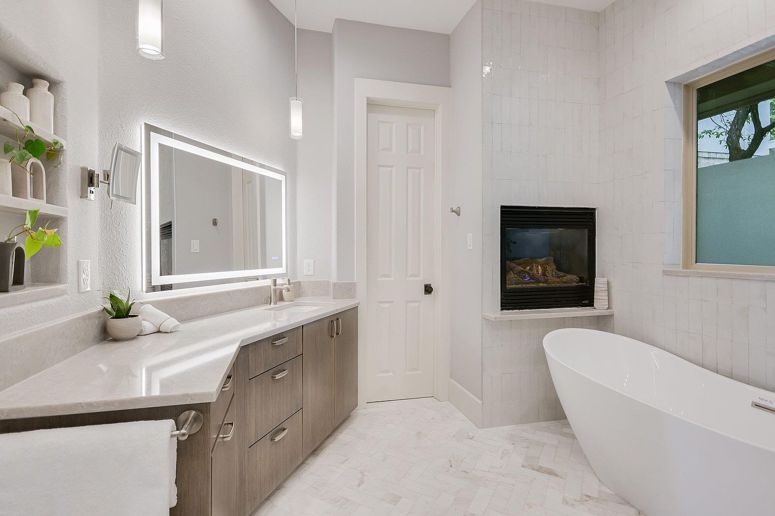 This is a well-lit primary bathroom featuring a modern design with a double vanity, a large illuminated mirror, and a freestanding bathtub. The walls are tiled, and there is a fireplace next to the tub, adding a luxurious touch. The color palette is primarily white and gray, creating a clean and serene atmosphere.