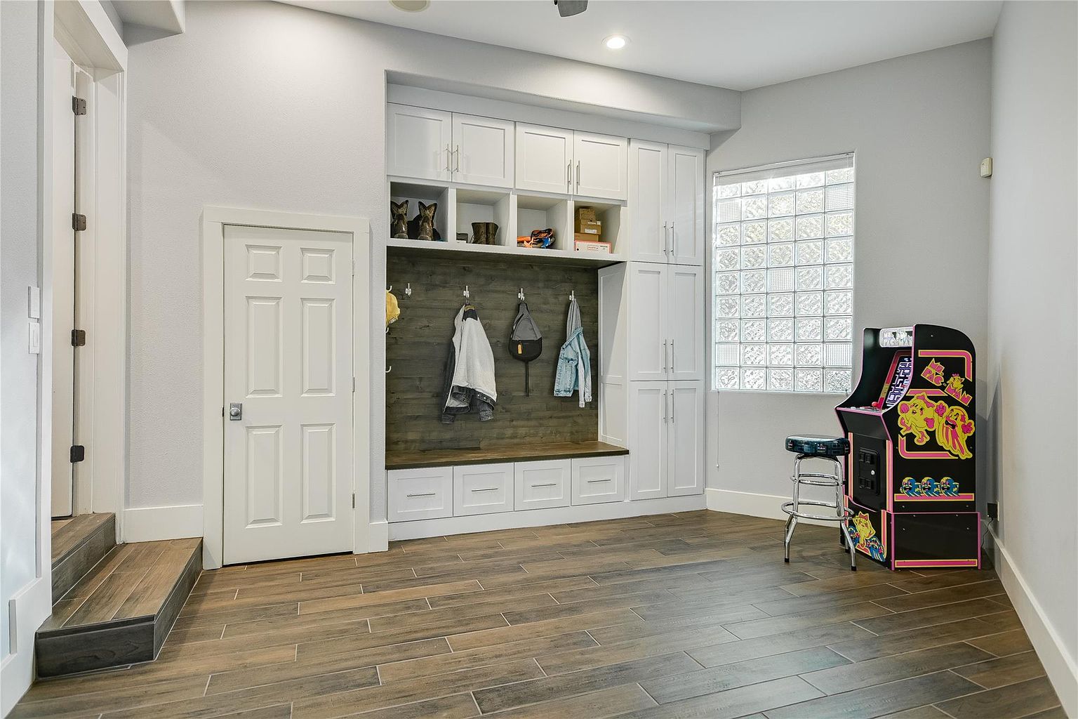 This interior shot showcases a well-organized laundry room or mudroom area. It features custom built-in cabinetry with a bench and coat hooks, providing ample storage. An arcade game adds a playful touch to the space, which is illuminated by natural light from a glass block window.
