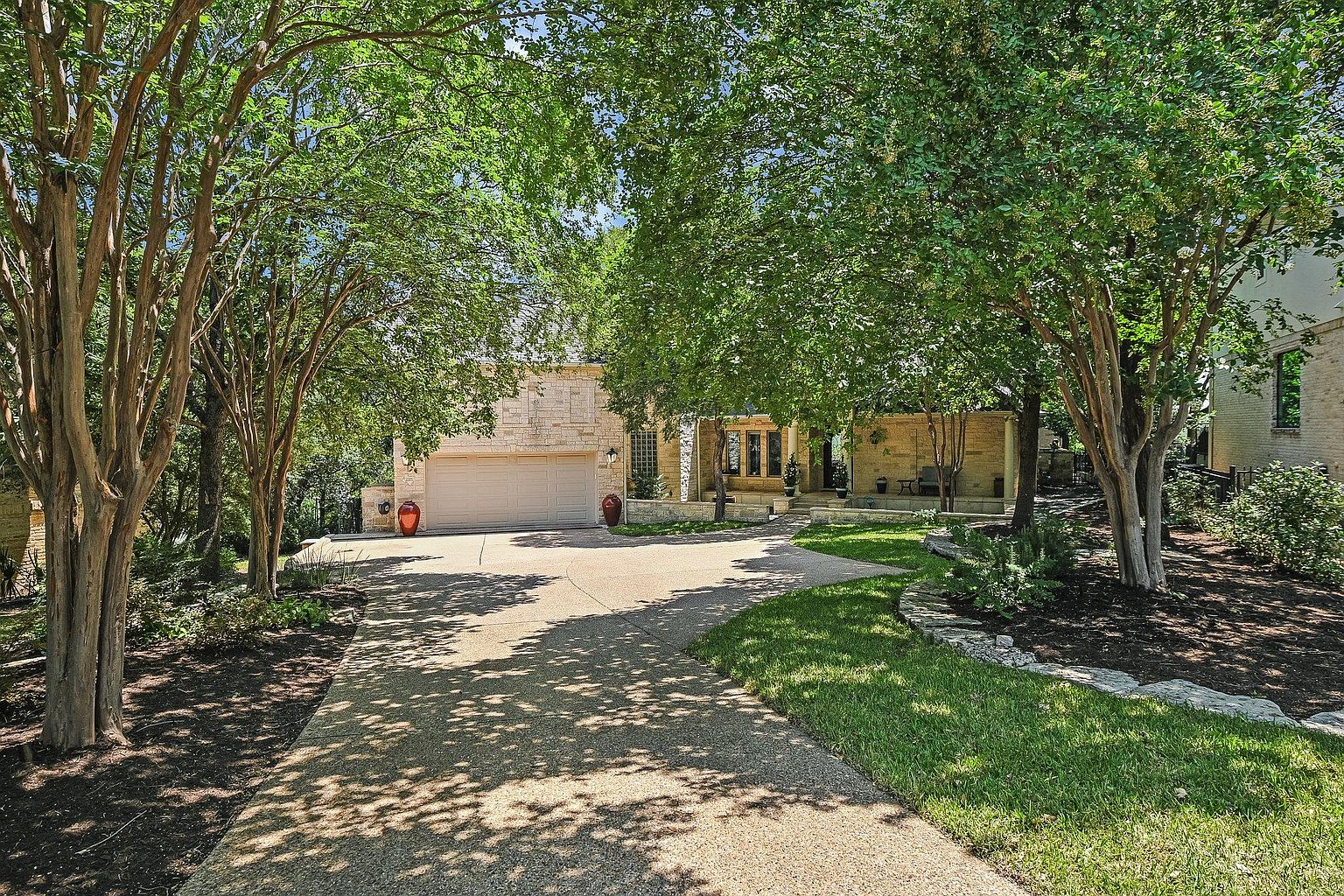 This is a front view of a single-story stone house with a two-car garage. The house is partially obscured by mature trees, creating a sense of privacy and shade. A long driveway leads up to the garage, and there is well-maintained landscaping with grass and mulch beds.