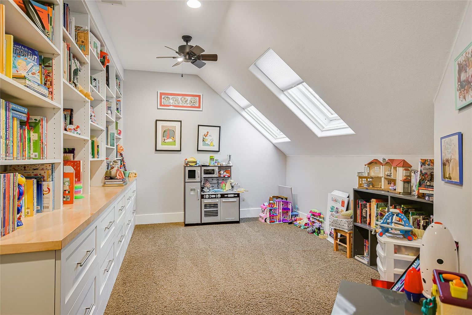 This is a charming children's playroom or bedroom featuring built-in bookshelves filled with books and toys. The room has a cozy feel with a neutral carpet, and natural light streams in through two skylights. A play kitchen and various toys are scattered around, creating a playful and inviting atmosphere.
