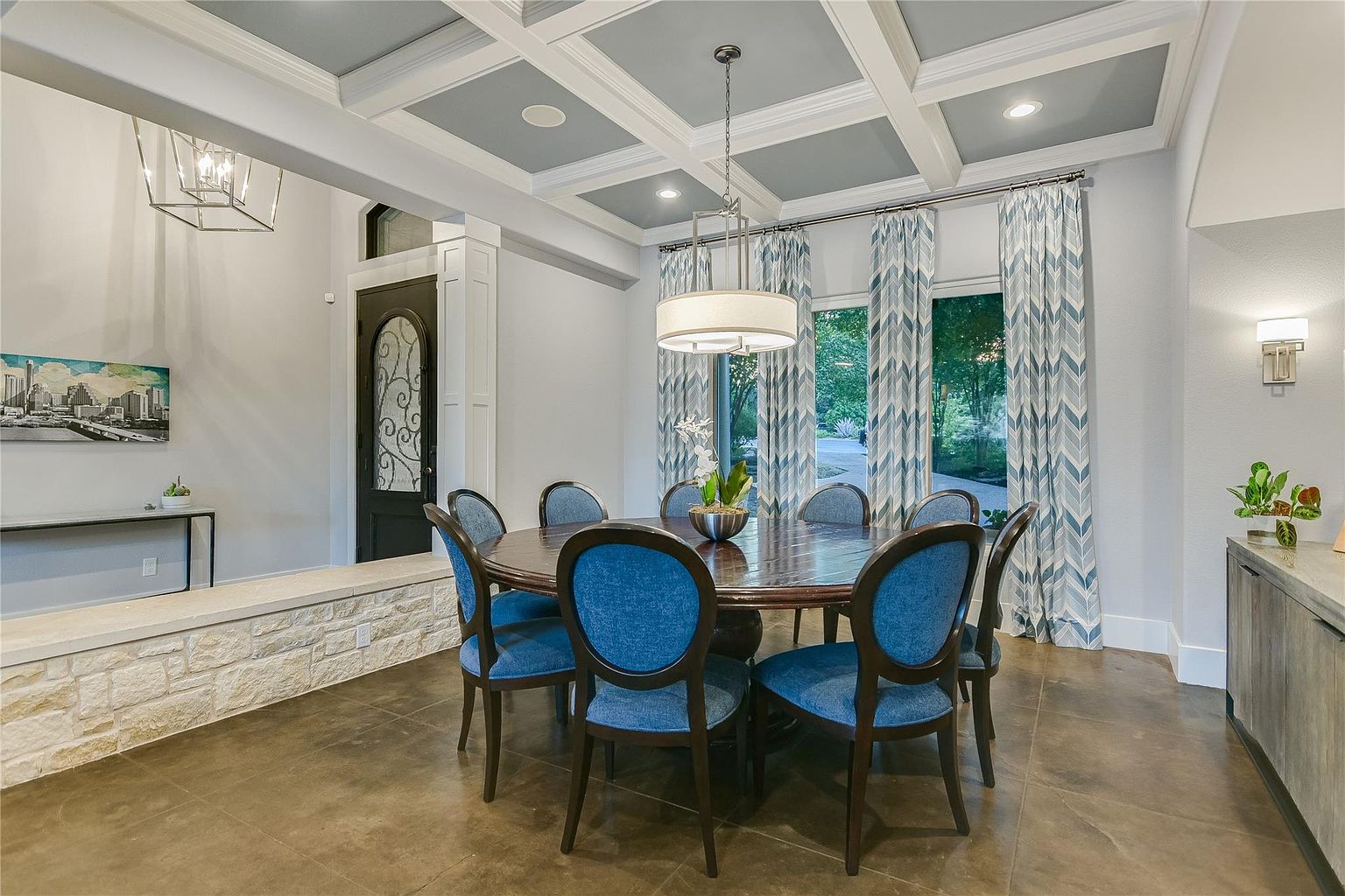 This is an interior shot of a dining room featuring a round wooden table with blue upholstered chairs. The room has a coffered ceiling with recessed lighting and a modern chandelier. Natural light streams in through the windows, which are adorned with patterned curtains, and there is a decorative stone accent wall near the entryway.