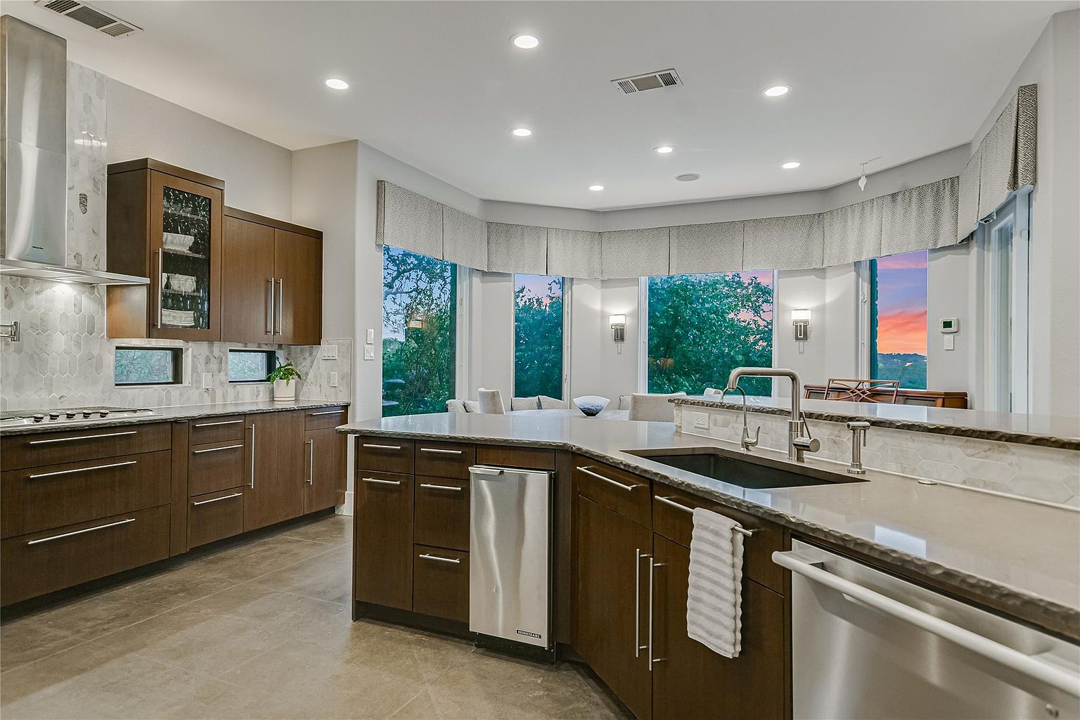 This is a spacious and modern kitchen featuring dark wood cabinetry with sleek, silver hardware. The countertops are light-colored stone, and stainless steel appliances add a contemporary touch. Large windows provide ample natural light and views of the outdoors, enhancing the open and airy feel of the space.