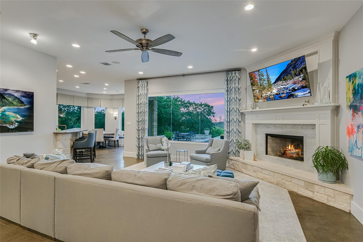 This is an interior shot of a well-lit living room featuring a large sectional sofa, a stone fireplace with a mounted television, and a large window offering a view of the outdoors. The room is decorated in neutral tones with pops of color from artwork and greenery, creating a comfortable and inviting atmosphere. The open floor plan connects to a dining area, enhancing the sense of spaciousness.