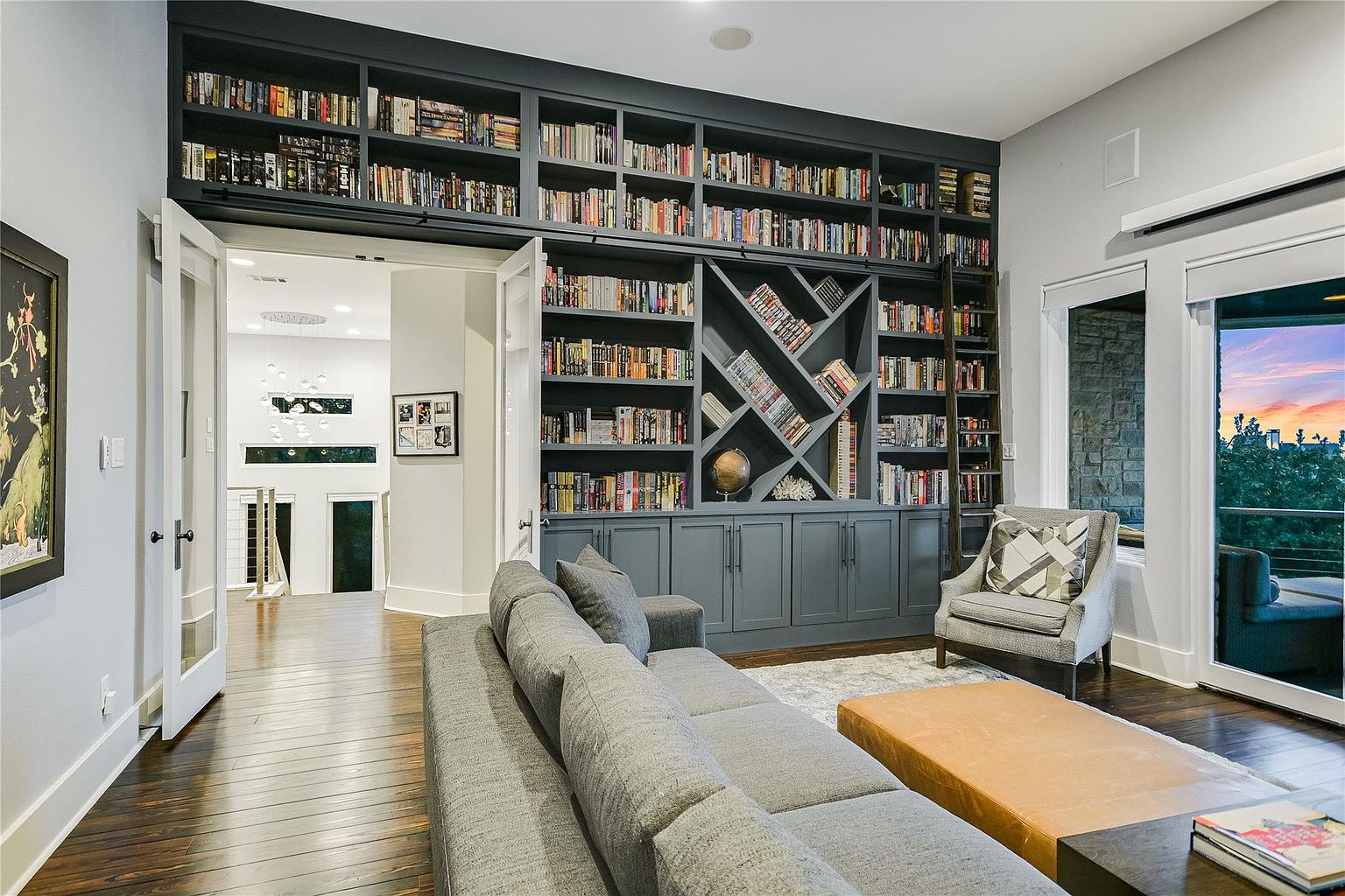This is an interior shot of a living room featuring a large, built-in bookcase filled with books, creating a cozy and sophisticated atmosphere. A gray sectional sofa and an armchair provide comfortable seating, while a large window offers natural light and a glimpse of the outdoors. The hardwood floors and neutral color palette contribute to a modern and inviting space.