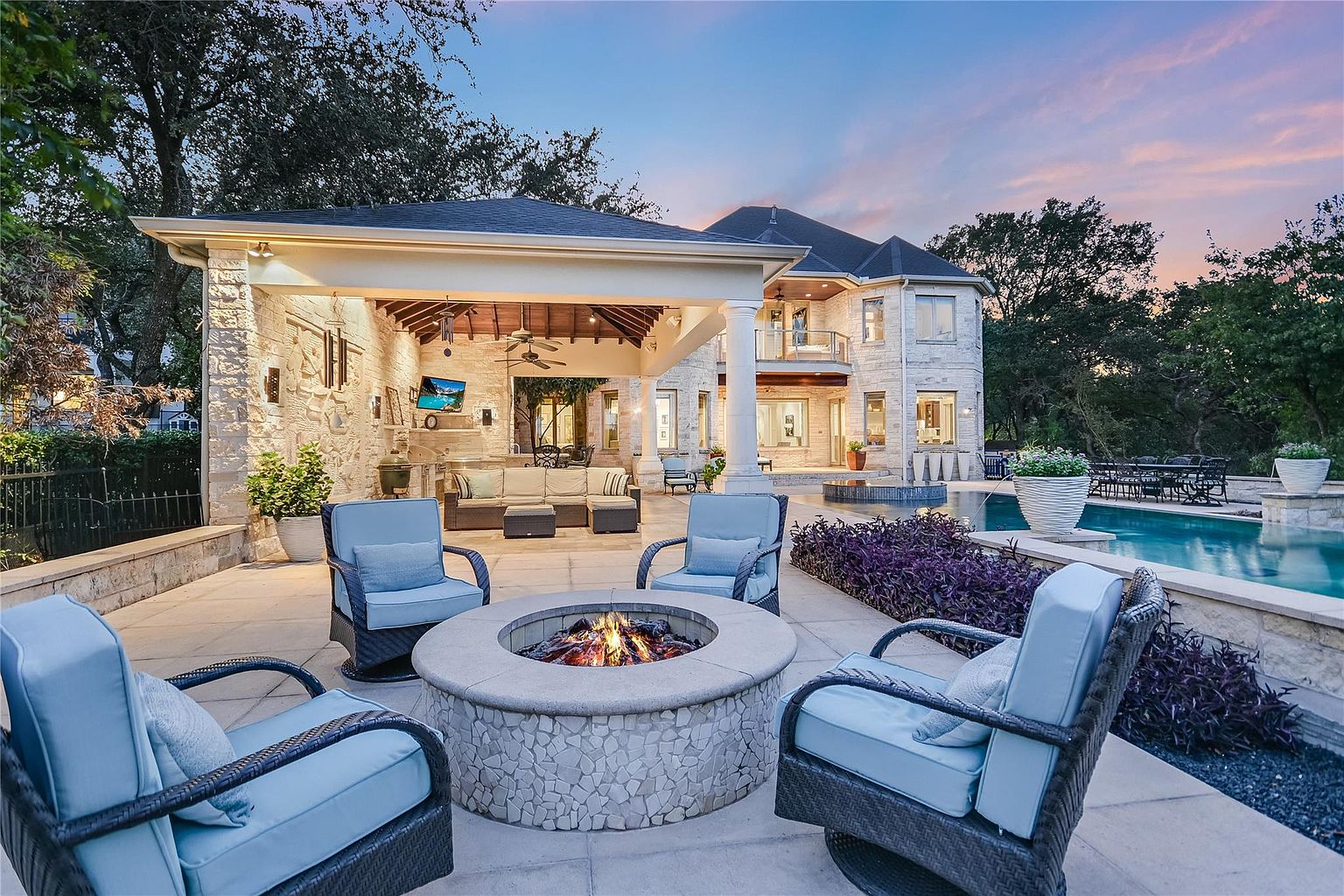 This image showcases a luxurious outdoor patio area, featuring a stone fire pit surrounded by comfortable blue cushioned chairs. The patio is part of a large, elegant stone house with a covered outdoor living space, complete with a television and ample seating. A swimming pool is visible in the background, adding to the property's appeal as an entertainment and relaxation haven.